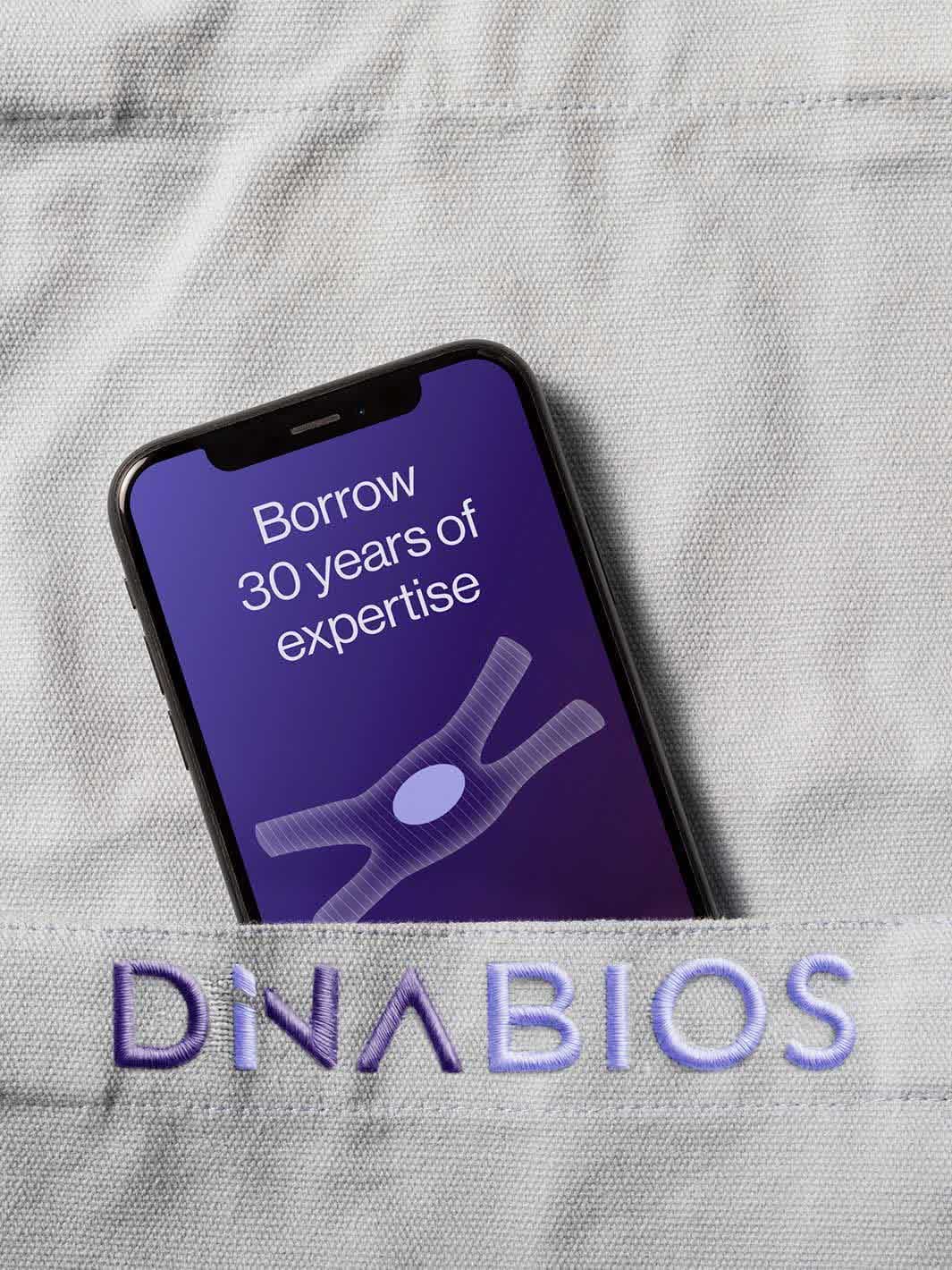 Phone in the pocket of a lab coat embroidered with the DiNABIOS logo, the screen says “Borrow 30 years of expertise”
