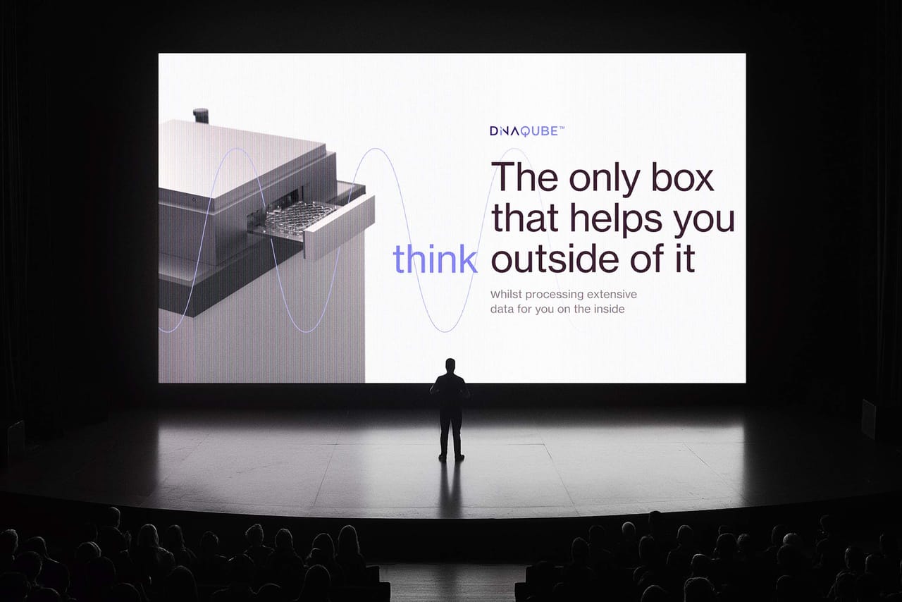 A speaker on stage in front of a slide: the DiNAQUBE, “The only box that helps you think outside of it”