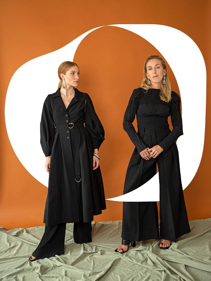 The two founders in black in front of an orange backdrop with a large black signet shape behind them
