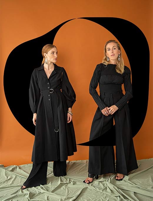 The two founders in black in front of an orange backdrop with a black signet shape