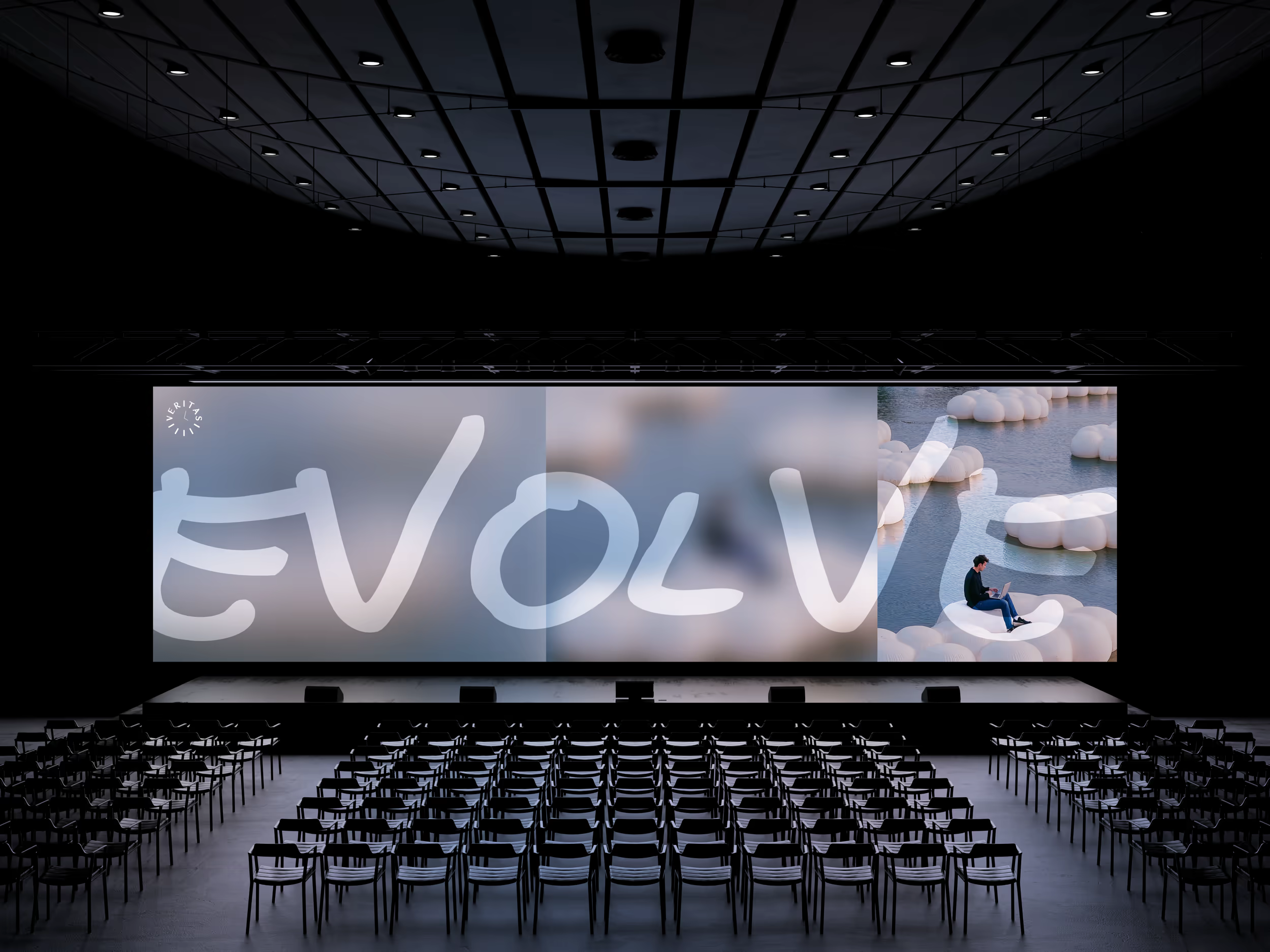 Dark event hall with a large screen showing the word “EVOLVE” and rows of chairs